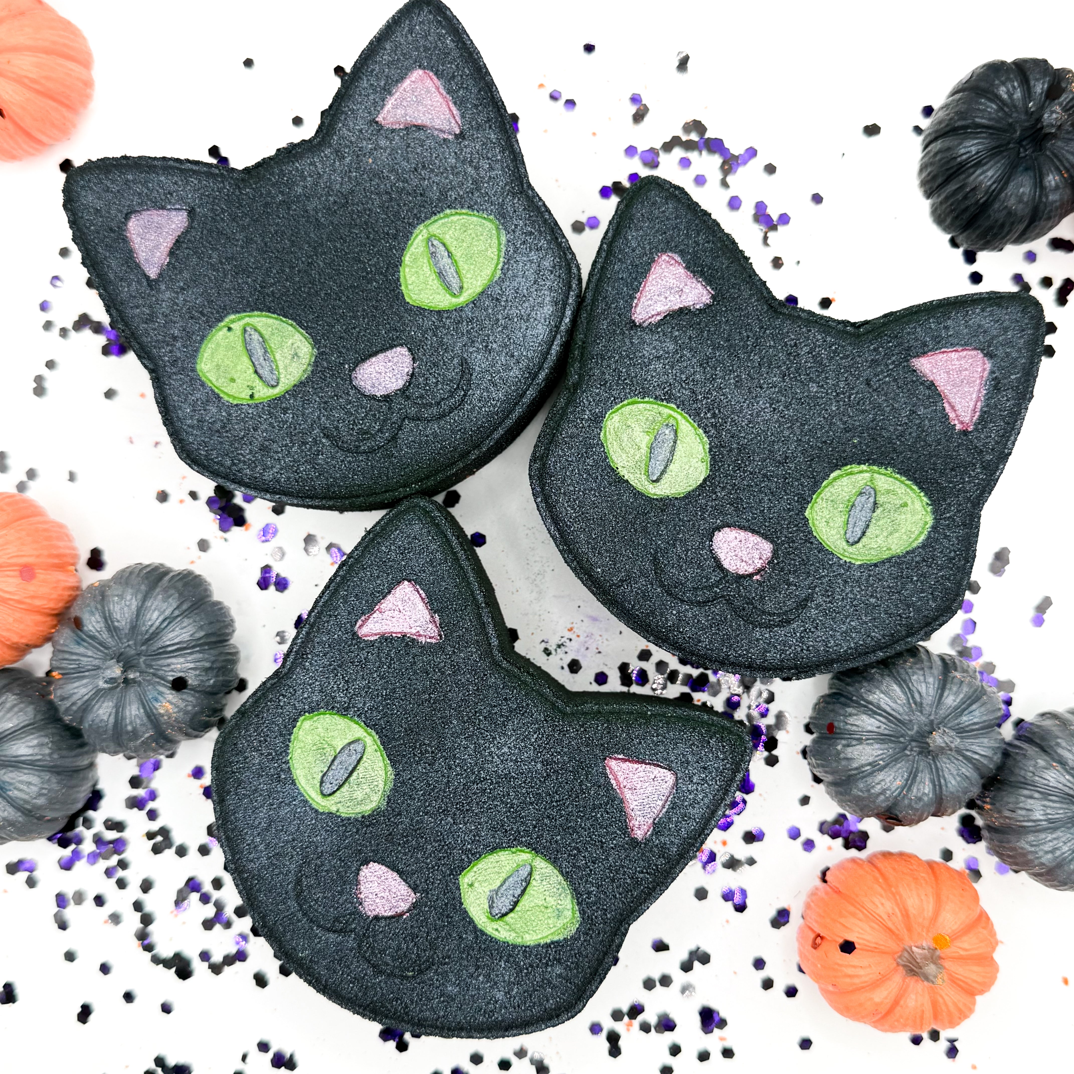 Three black cat bath bombs with green eyes, pink noses, and pink ears, arranged on a white background with small pumpkins and purple sprinkles.
