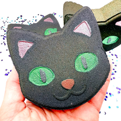 Hand holding Rusted Acre Soap Co.’s Black Cat Bath Bomb with green eyes and pink ears — a spooky, kid-friendly Halloween bath treat.