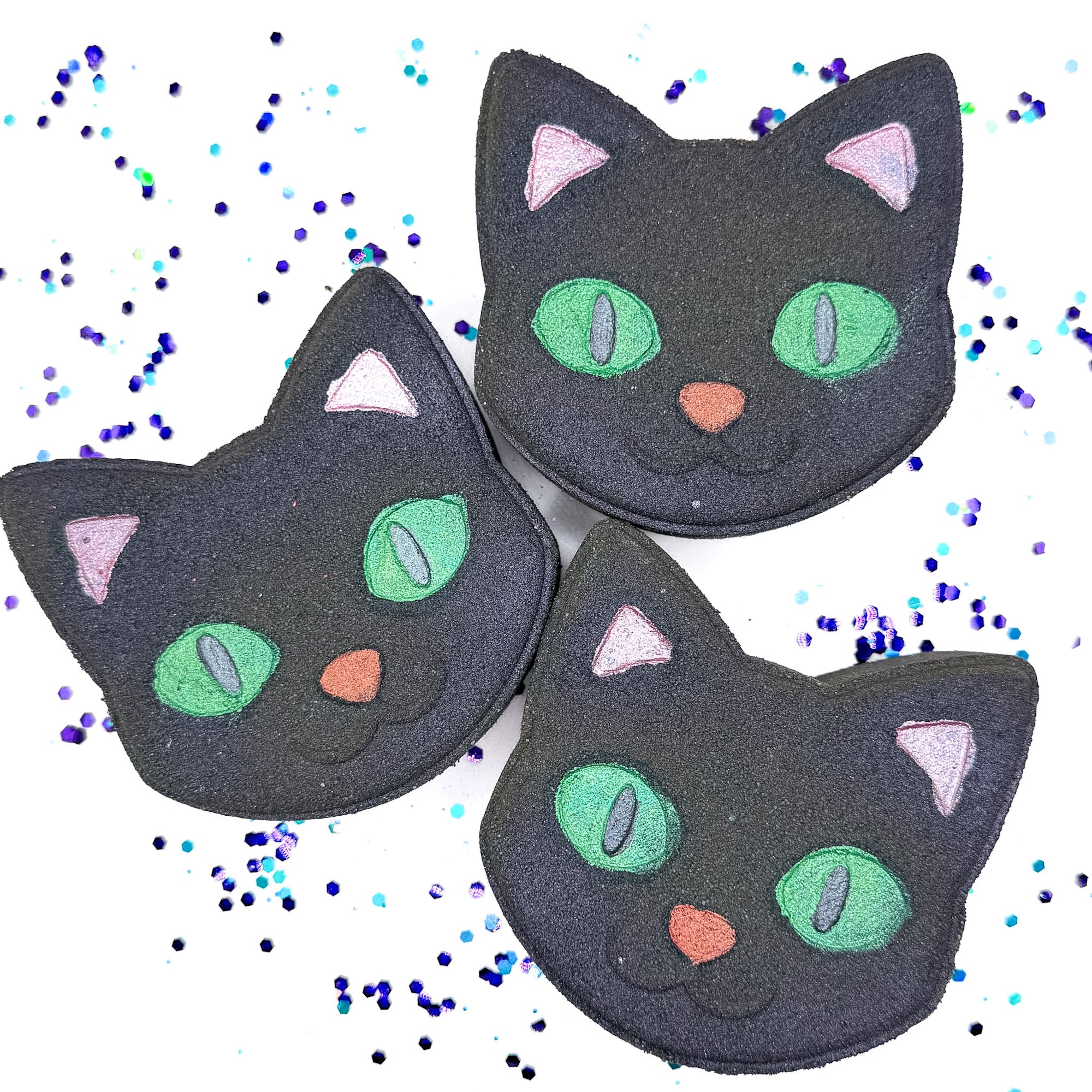 Three handmade Black Cat Bath Bombs from Rusted Acre Soap Co., each shaped like a cat’s face with green eyes, pink ears, and a red or pink nose, displayed on a white background with scattered purple and teal confetti