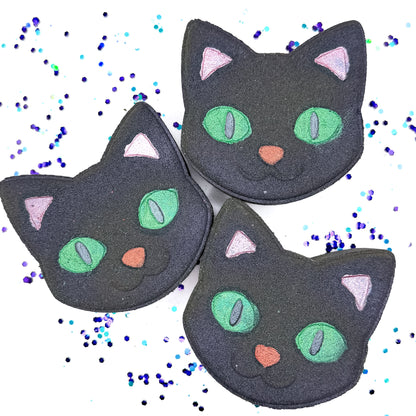 Three handmade Black Cat Bath Bombs from Rusted Acre Soap Co., each shaped like a cat’s face with green eyes, pink ears, and a red or pink nose, displayed on a white background with scattered purple and teal confetti
