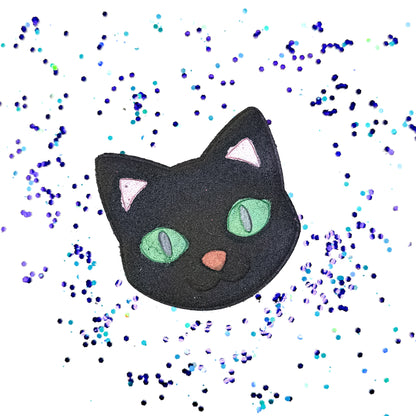 Rusted Acre Soap Co.‘s handmade Black Cat Bath Bomb shaped like a cat’s face with green eyes, pink ears, and a soft pink nose, surrounded by scattered blue and purple bath confetti on a white background.
