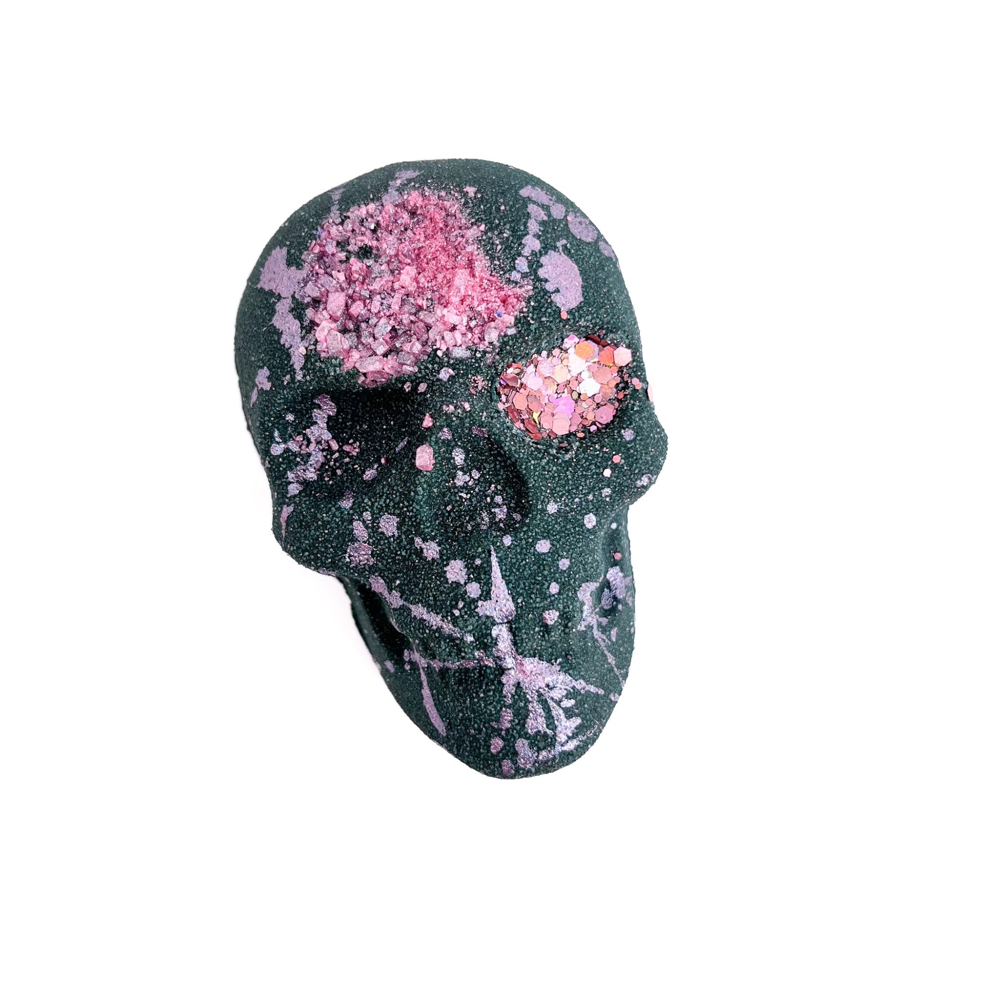 Black skull shaped bath bomb splattered with light pink mica. Pink sea salt is embedded in the forehead with gold mica hand-painted around edges.