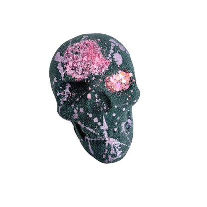 Black skull shaped bath bomb splattered with light pink mica. Pink sea salt is embedded in the forehead with gold mica hand-painted around edges.
