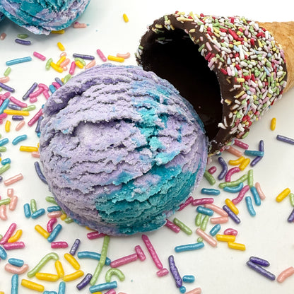 One purple and blue mermaid kiss scented bubble bath scoop handmade by Rusted Acre Soap Co. Scoop looks like an ice cream scoop. White background with sprinkles and ice cream cone prop on a white background. 