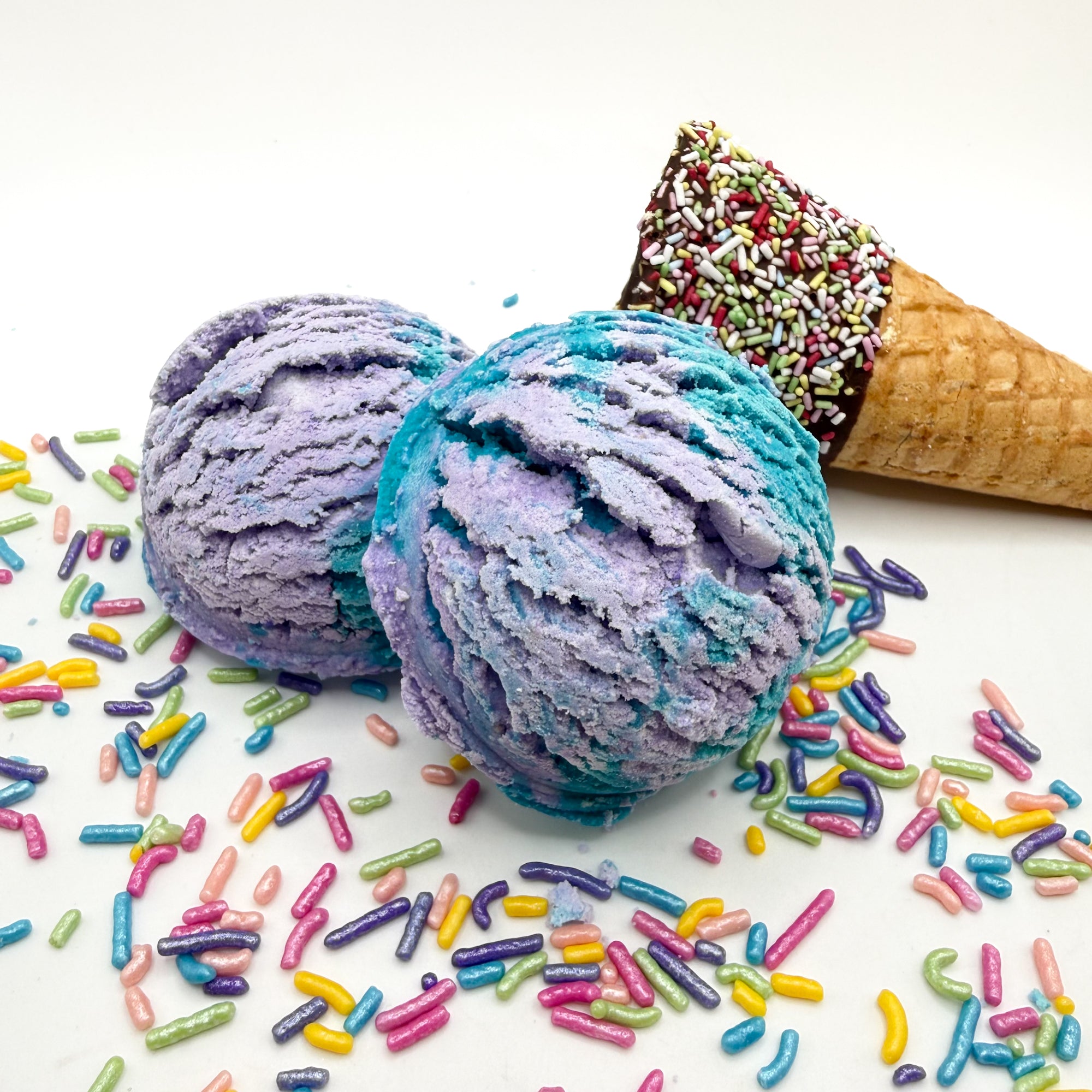 Two purple and blue mermaid kiss scented bubble bath scoop handmade by Rusted Acre Soap Co. Scoop looks like an ice cream scoop. White background with sprinkles and ice cream cone prop on a white background. 