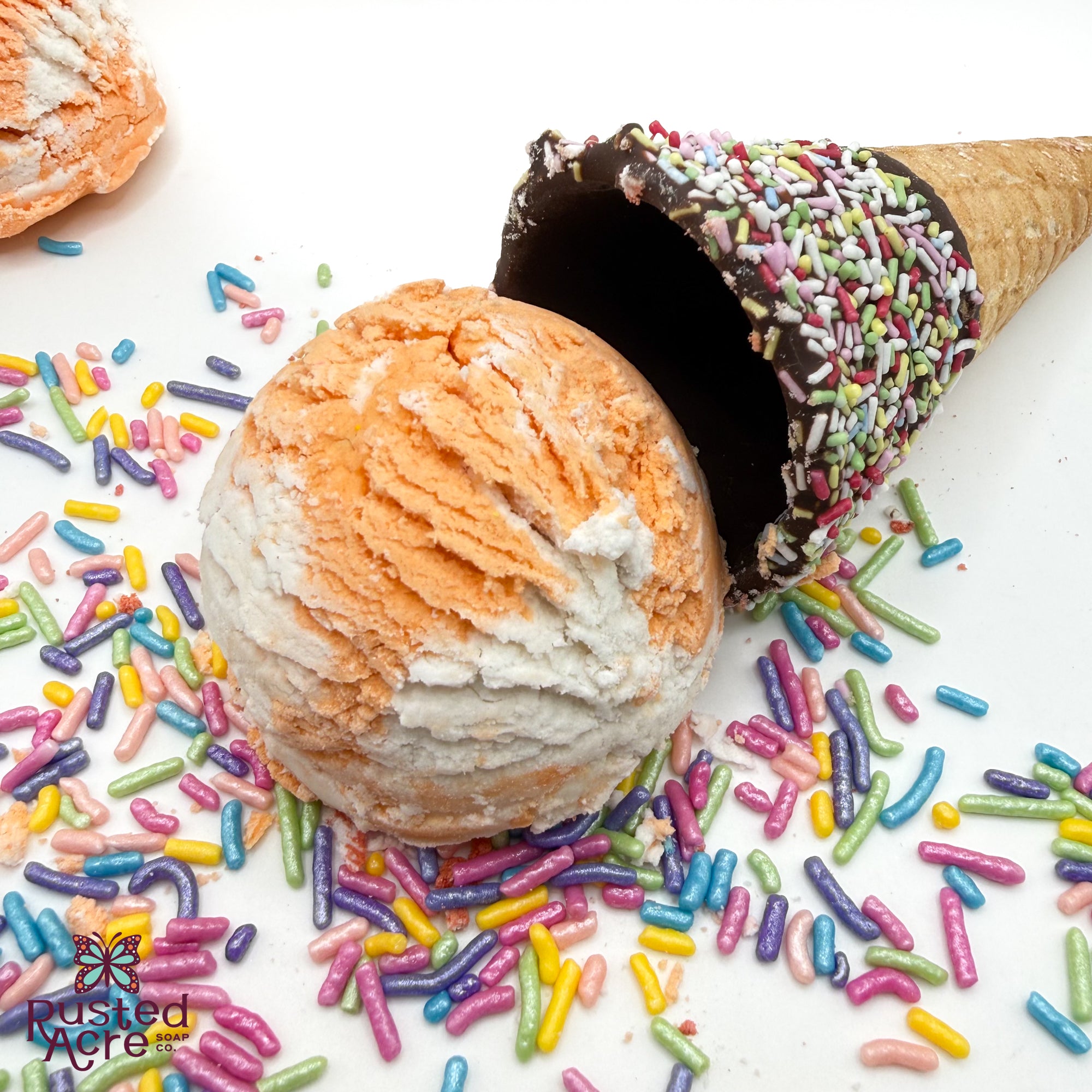 One orange and white orange creamcicle scented bubble bath scoop handmade by Rusted Acre Soap Co. Scoop looks like an ice cream scoop. White background with sprinkles and ice cream cone prop on a white background. 