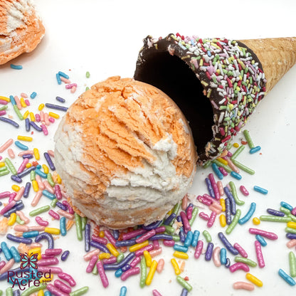 One orange and white orange creamcicle scented bubble bath scoop handmade by Rusted Acre Soap Co. Scoop looks like an ice cream scoop. White background with sprinkles and ice cream cone prop on a white background. 