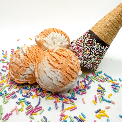 Three orange and white orange creamcicle scented bubble bath scoop handmade by Rusted Acre Soap Co. Scoop looks like an ice cream scoop. White background with sprinkles and ice cream cone prop on a white background. 