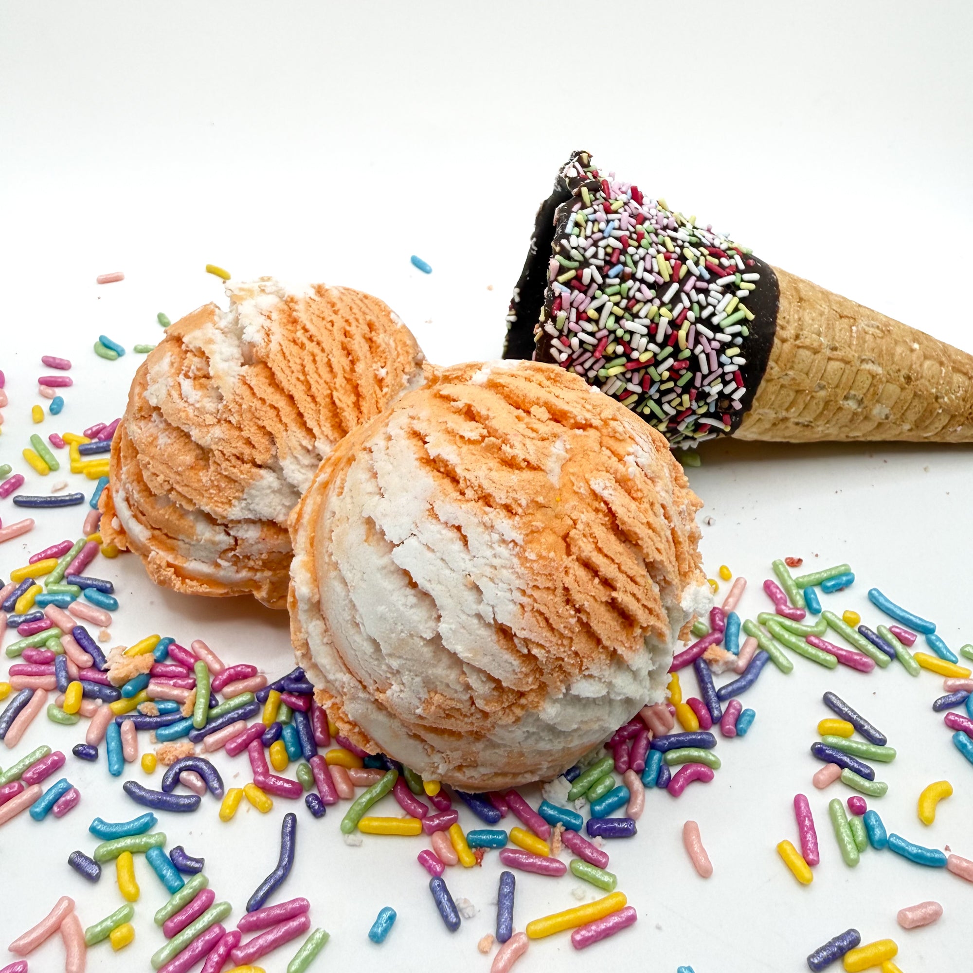 Two orange and white orange creamcicle scented bubble bath scoop handmade by Rusted Acre Soap Co. Scoop looks like an ice cream scoop. White background with sprinkles and ice cream cone prop on a white background. 