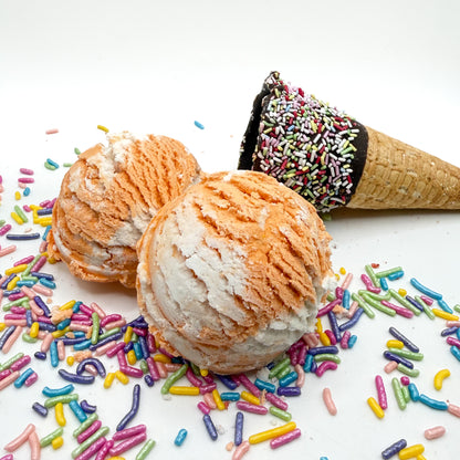 Two orange and white orange creamcicle scented bubble bath scoop handmade by Rusted Acre Soap Co. Scoop looks like an ice cream scoop. White background with sprinkles and ice cream cone prop on a white background. 