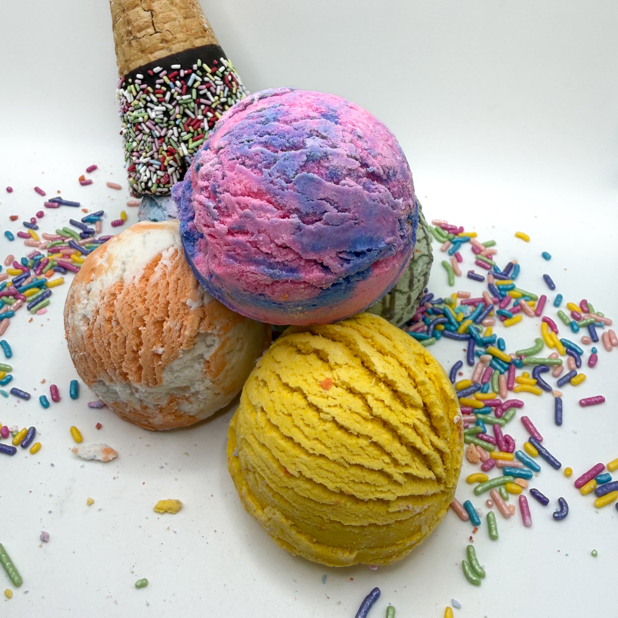 Multiple bubble bath scoops handmade by Rusted Acre Soap Co. Scoop looks like an ice cream scoop. White background with sprinkles and ice cream cone prop on a white background. 
