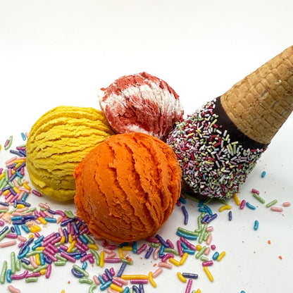 Three different colored and scented bubble bath scoops handmade by Rusted Acre Soap Co. Scoop looks like an ice cream scoop and are orange, yellow and red and white. White background with sprinkles and ice cream cone prop on a white background. 