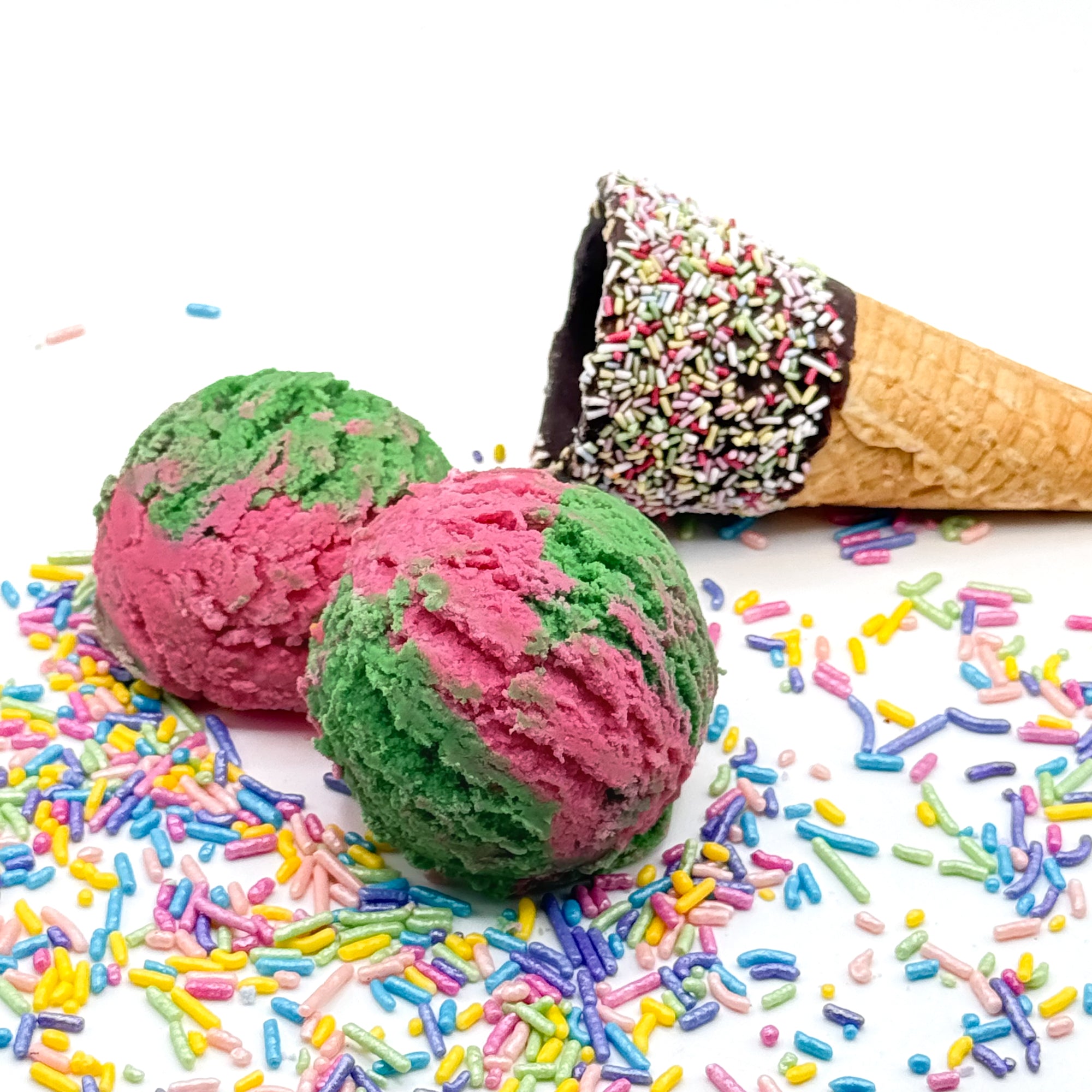 Two scoops of pink and green watermelon bubble bath scoops shaped like ice cream with a cone prop on a white background with colorful sprinkles.