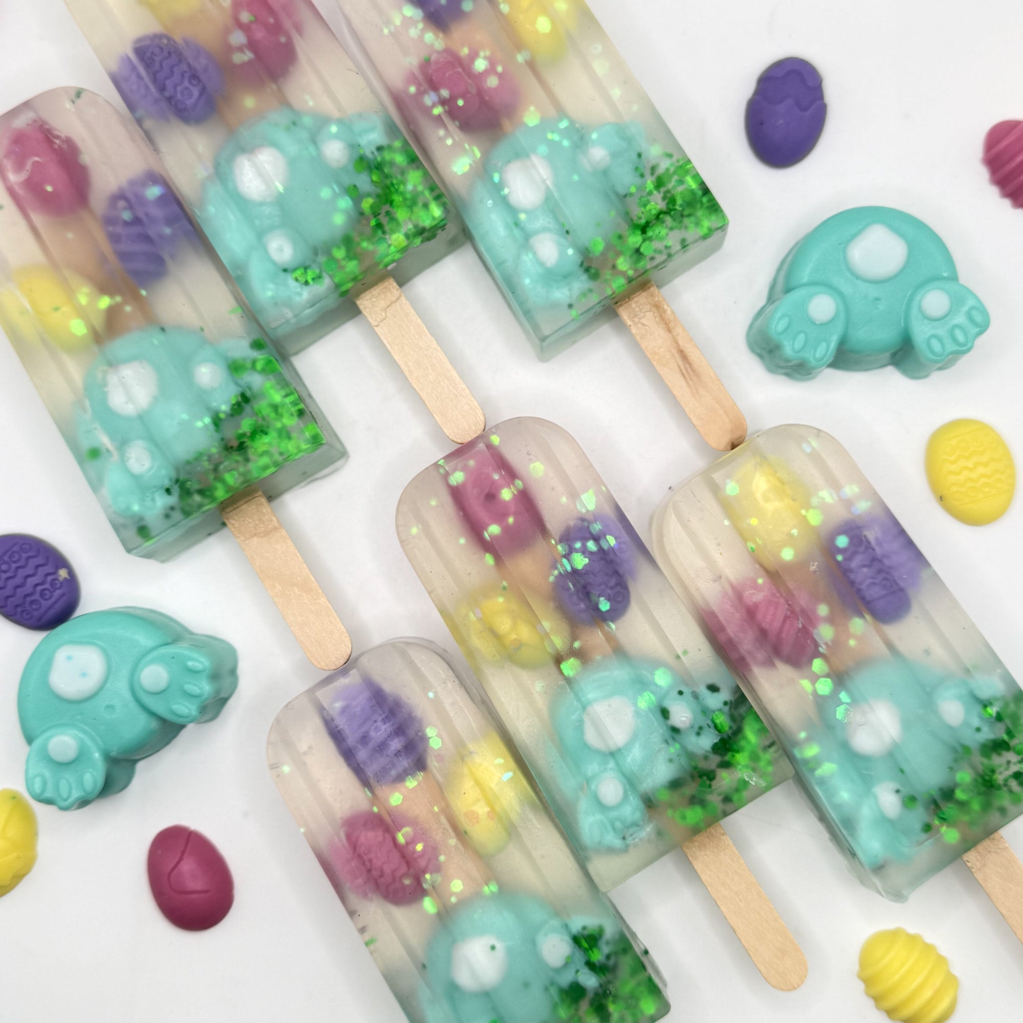 Six colorful Easter popsicle shaped soap bars with bunny and egg soap embedded inside on a white background. Handmade by Rusted Acre Soap Co.