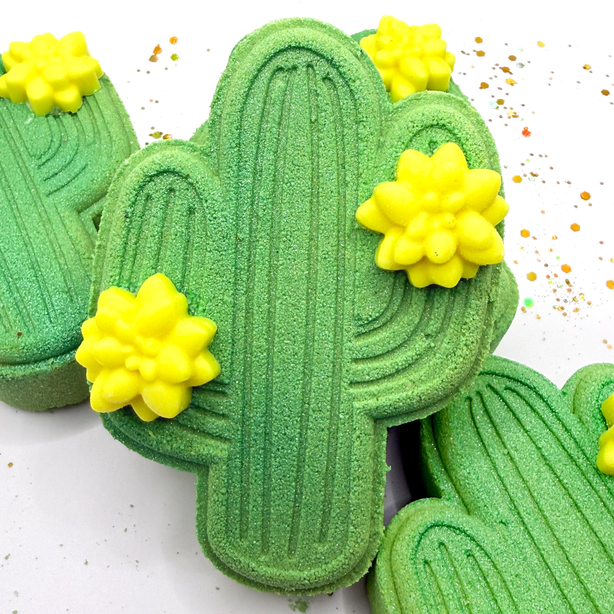 One green cactus shaped bath bomb standing up with other lying. Each adorned with two 
 light yellow cactus flower mini soap.