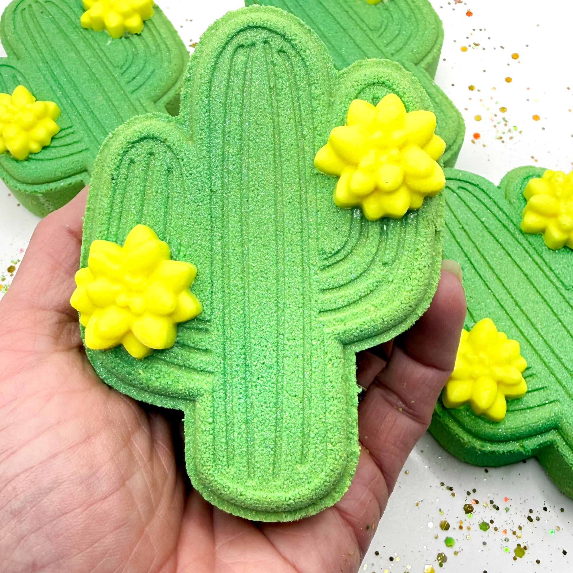 Green cactus shaped bath bomb with yellow flowers held in a hand against a white background.