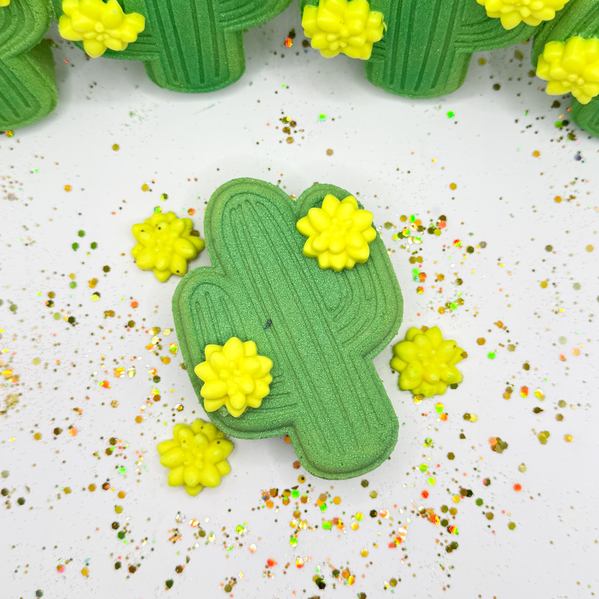 One green cactus shaped bath bombs standing with multiple cactus bath bombs in the background Each adorned with two 
 light yellow cactus flower mini soap.