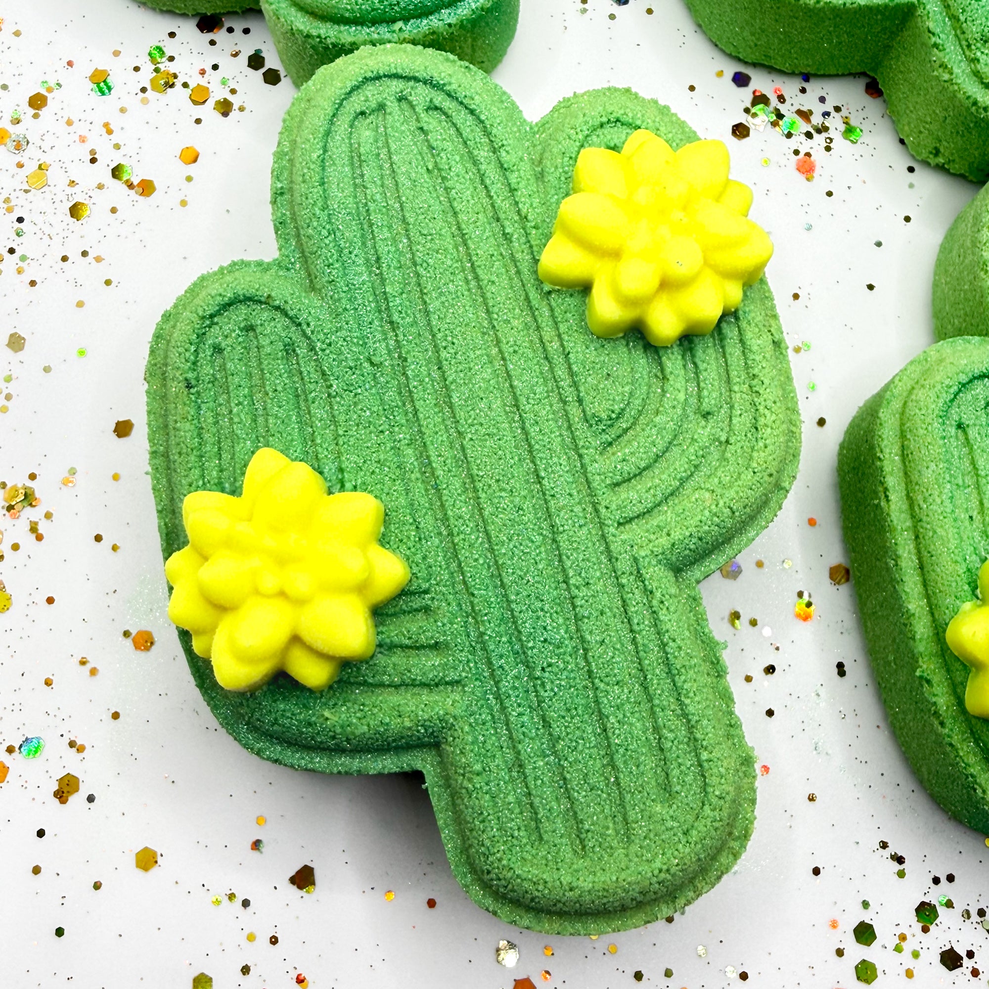 One green cactus shaped bath bombs. One standing two lying. Each adorned with two 
 light yellow cactus flower mini soap.