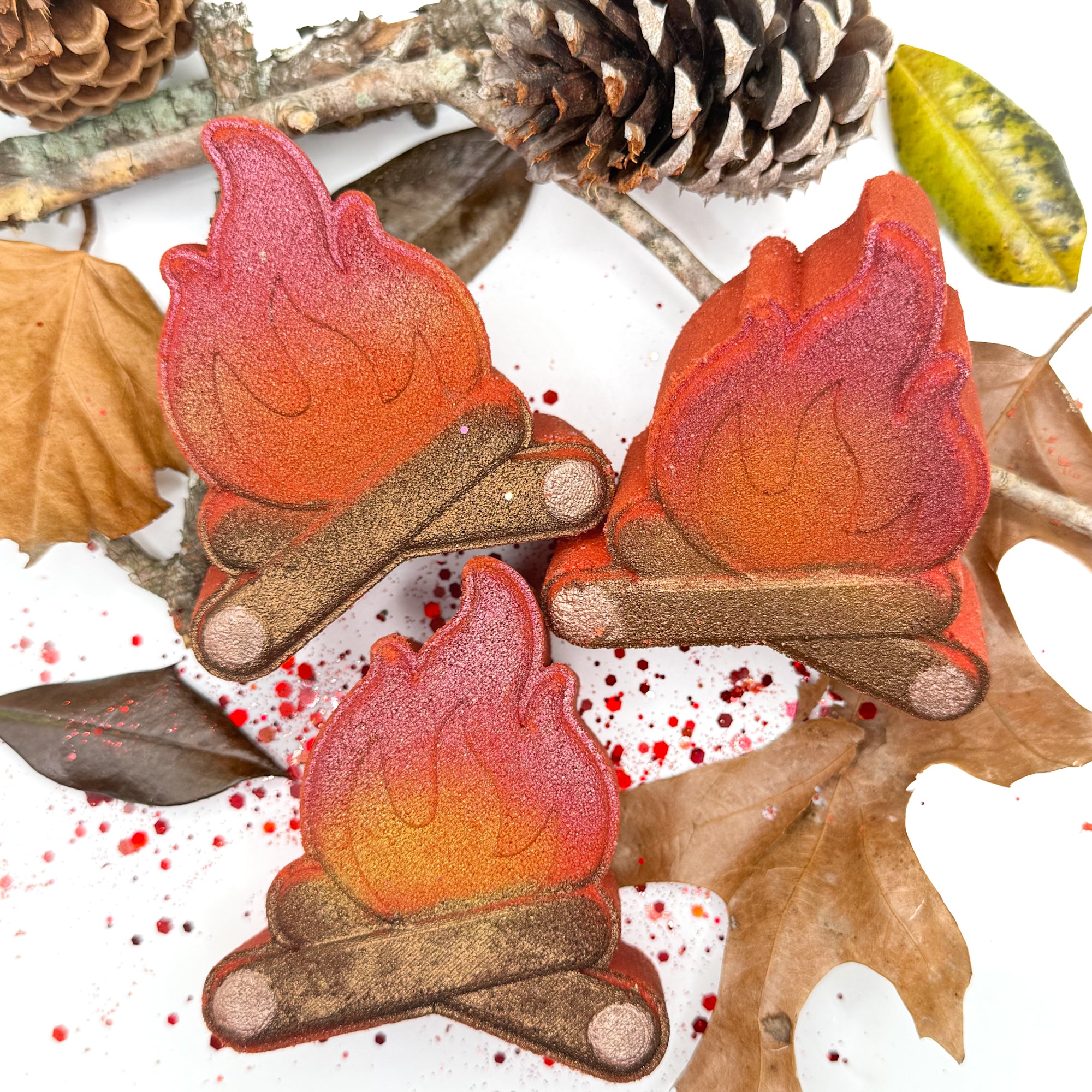 Three hand painted campfire bath bombs with leaves, sticks and pinecones on a white background.