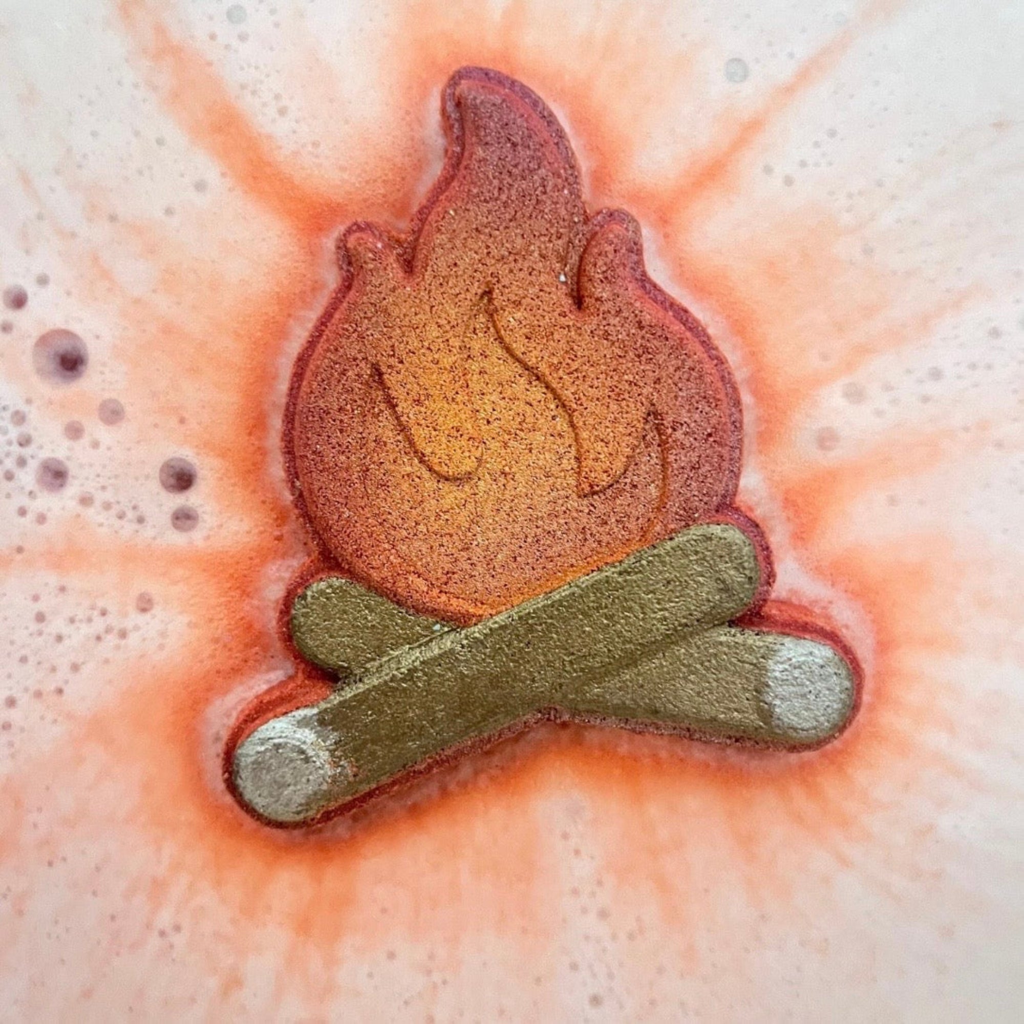 Handmade campfire bath bomb in the water with white, red, orange foam surrounding the bath bomb. 