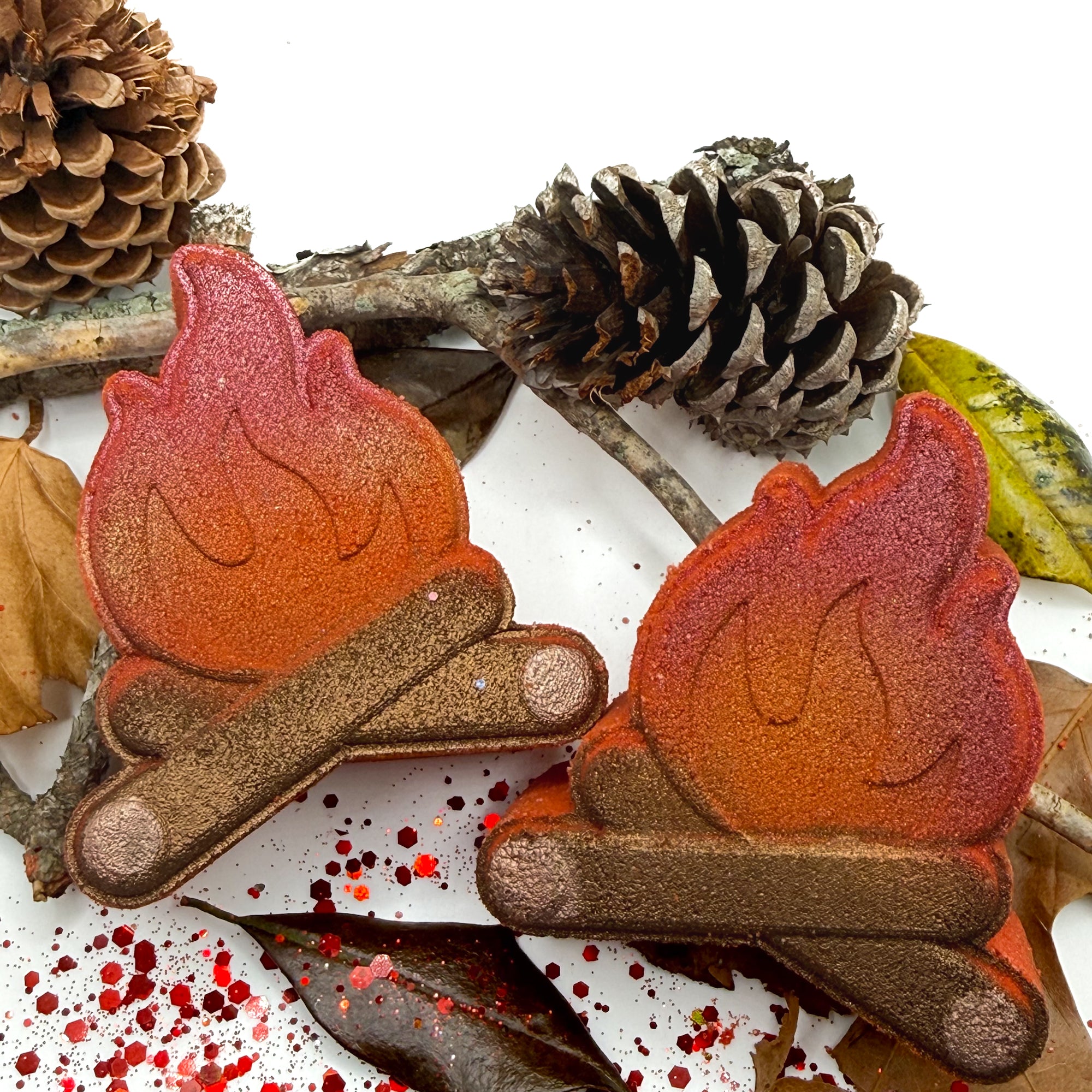 Two campfire shaped bath bombs with pine cones sticks, and leaves on a decorated white background.