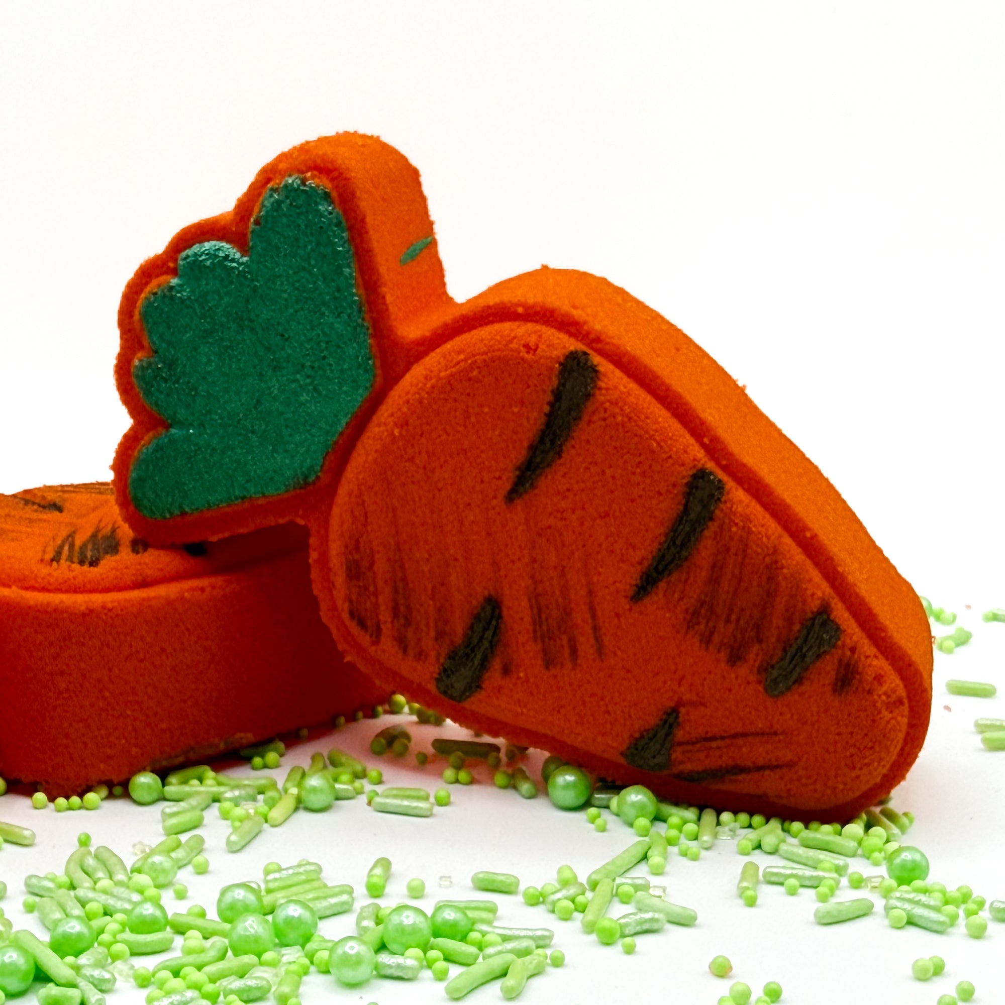 Front view of a hand painted orange Carrot  bath bomb with green tops and “dirt” paint on a white background. 