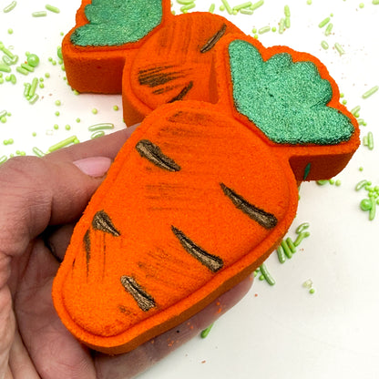 Handmade carrot bath bombs with green tops held by a hand on a white background with green sprinkles.