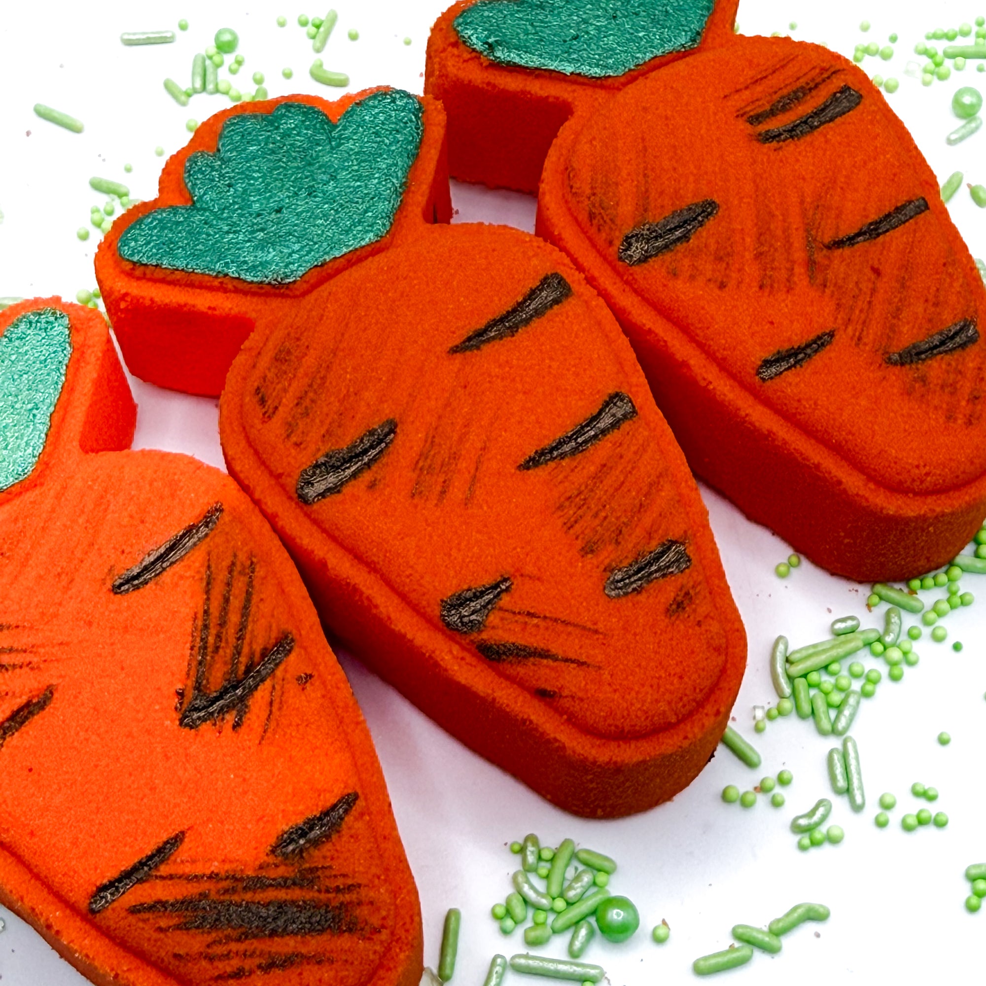 Three hand painted orange Carrot  bath bombs with green tops and “dirt” paint on a white background.