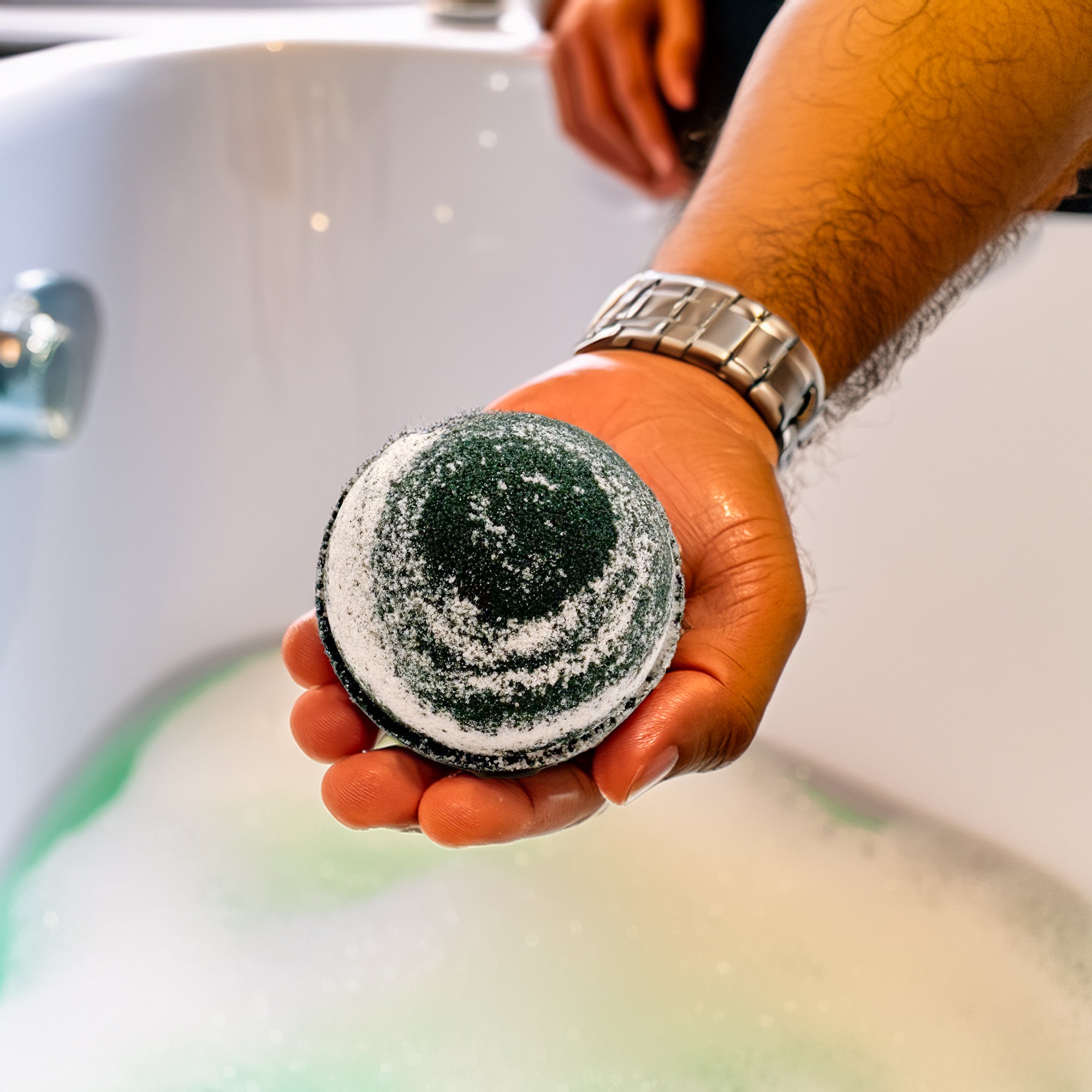 A handmade Cashmere bath bomb handmade by Rusted Acre Soap Co. in a hand over a bathtub.