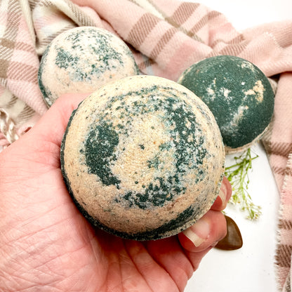 One tan and brown bath bomb held in a hand with two other bath bombs in the background. 