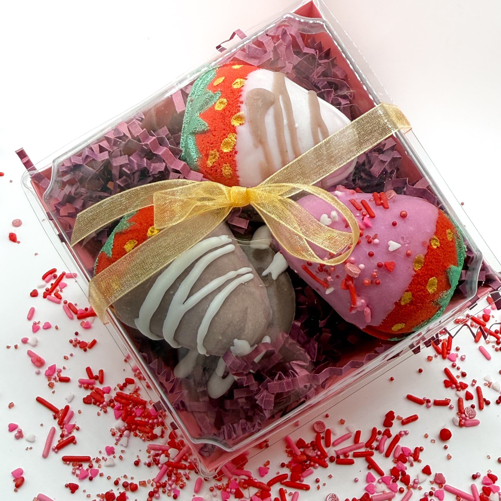 Multiple chocolate covered strawberry bath bombs hand made by Rusted Acre Soap Co with pink, white and brown chocolate hand dipped to look regular strawberries packaged in a gift box with a gold bow. 