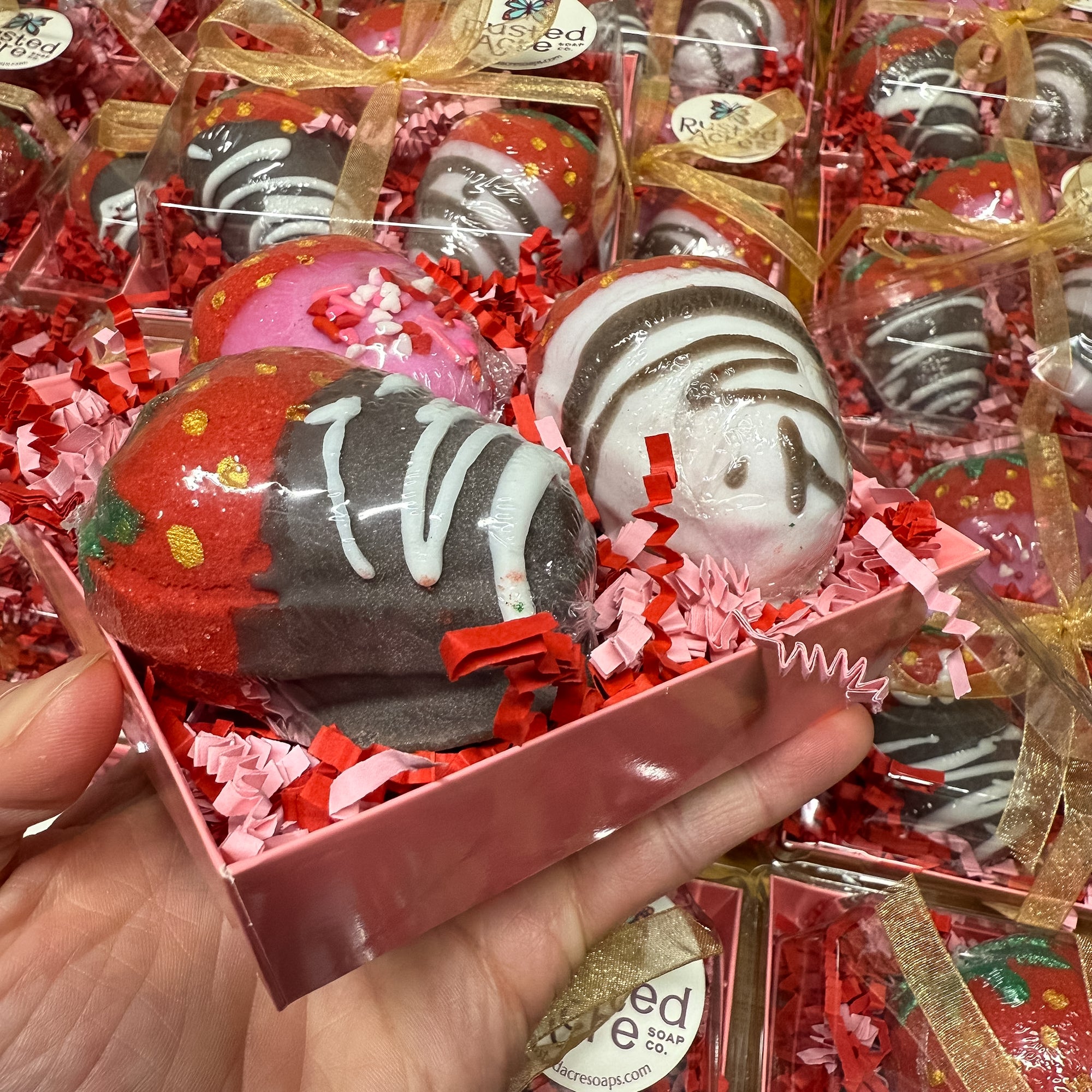Hand holding a box of decorated chocolate covered strawberry bath bombs in red and pink packaging.