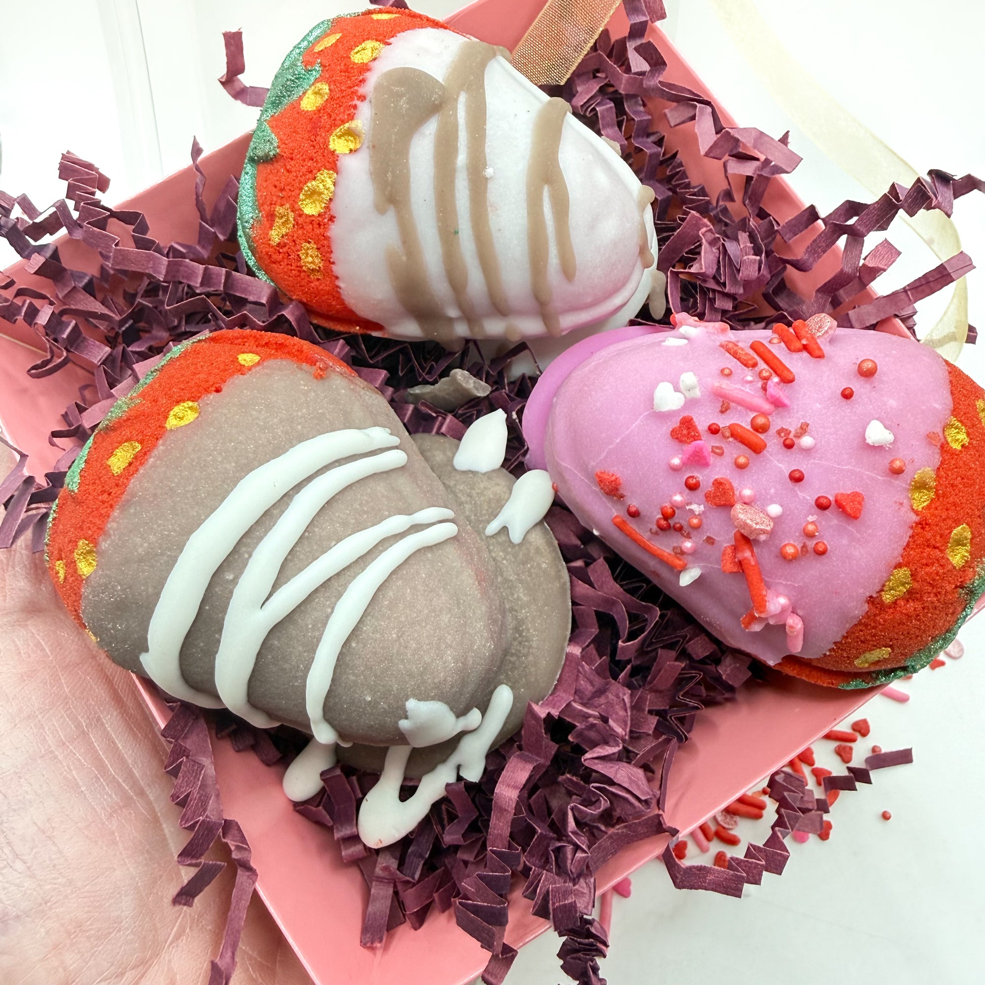 Three colorful chocolate covered strawberry bath bombs with decorative toppings in a pink box with burgundy shredded paper.