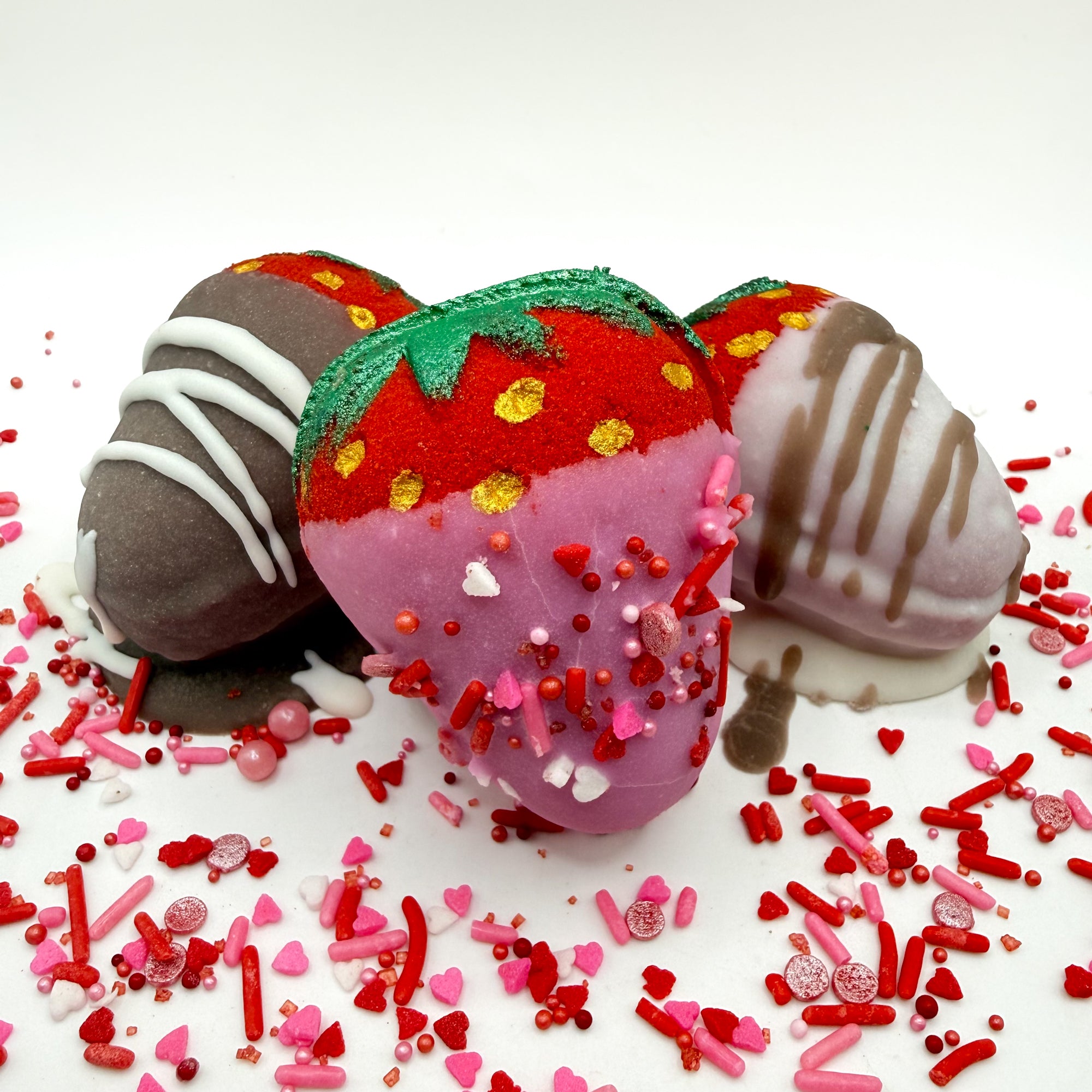 Three handmade chocolate covered strawberry bath bombs with  sprinkles and drizzle on a white background