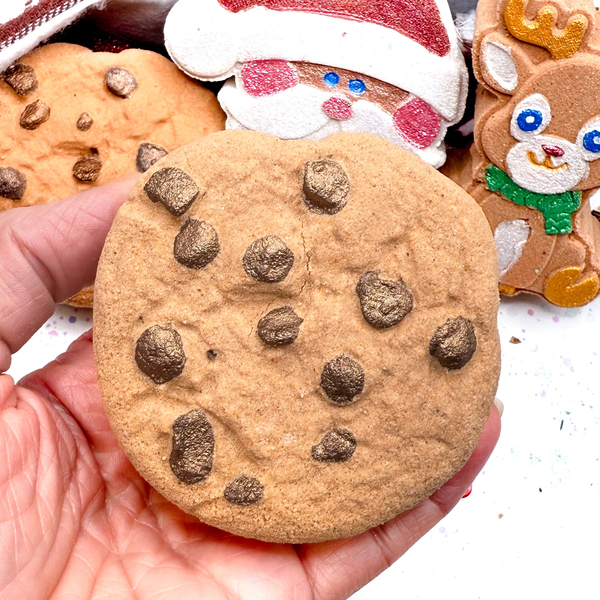 Hand holding a chocolate chip cookie bath bomb made by Rusted Acre Soap Co with other bath bombs in the background