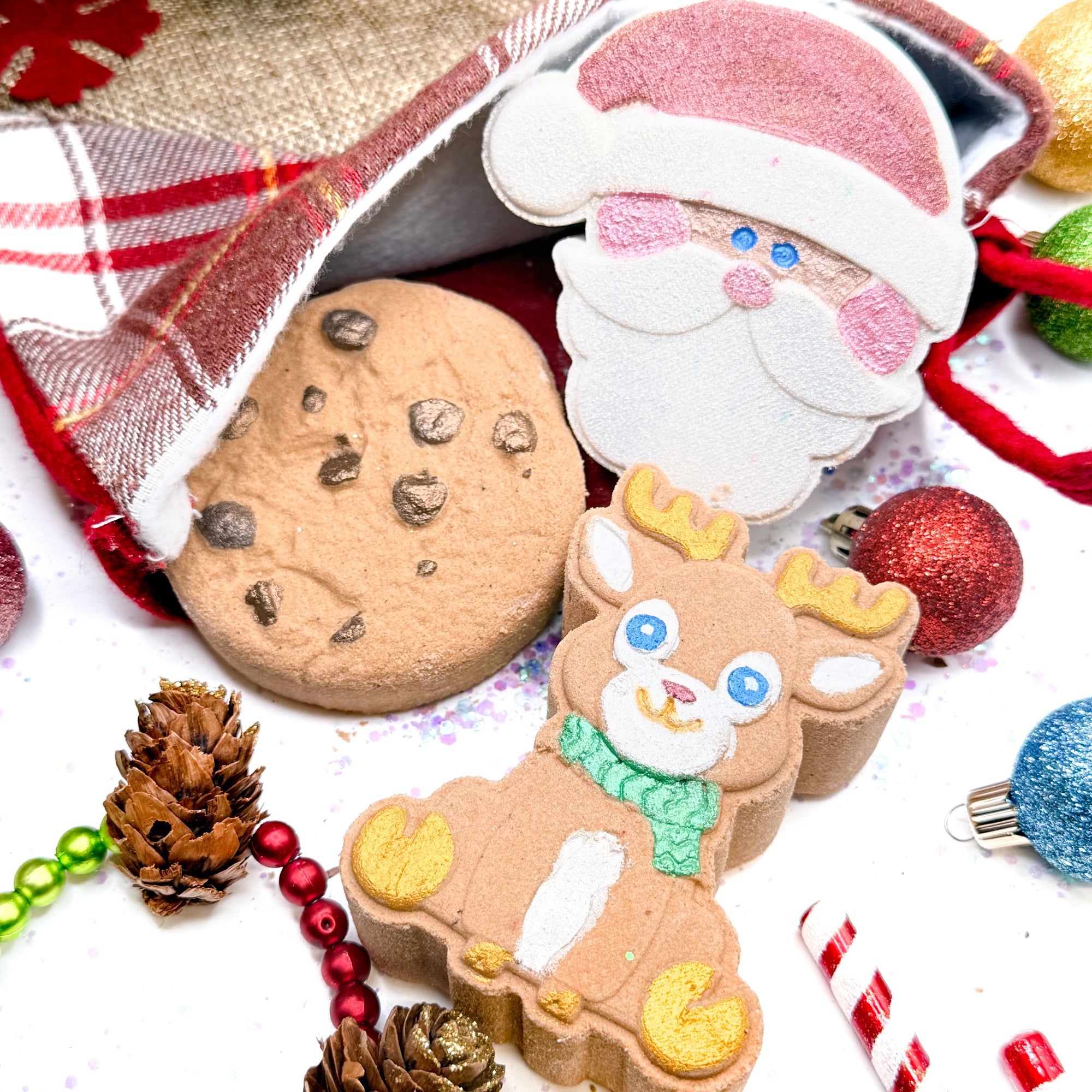 Christmas-themed bath bombs shaped like Santa Claus and a reindeer, surrounded by festive decorations on a white background.