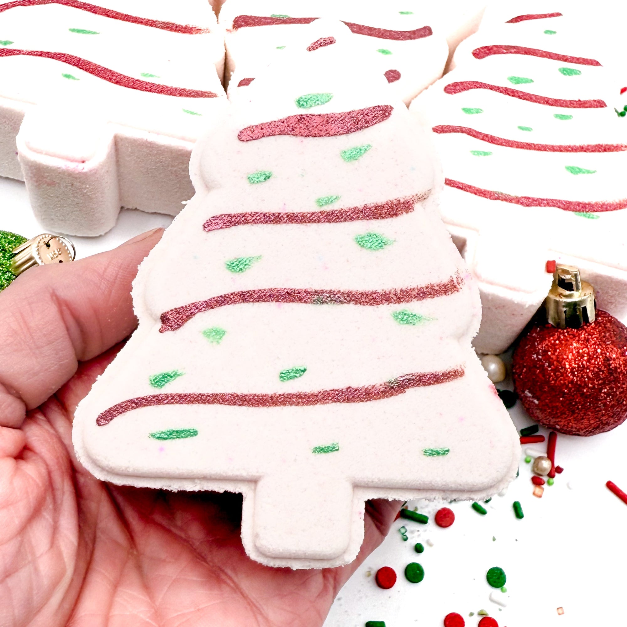 White Christmas tree shaped bathh bomb made by Rusted Acre Soap Co with red and green decorations held in a hand, surrounded by festive items.