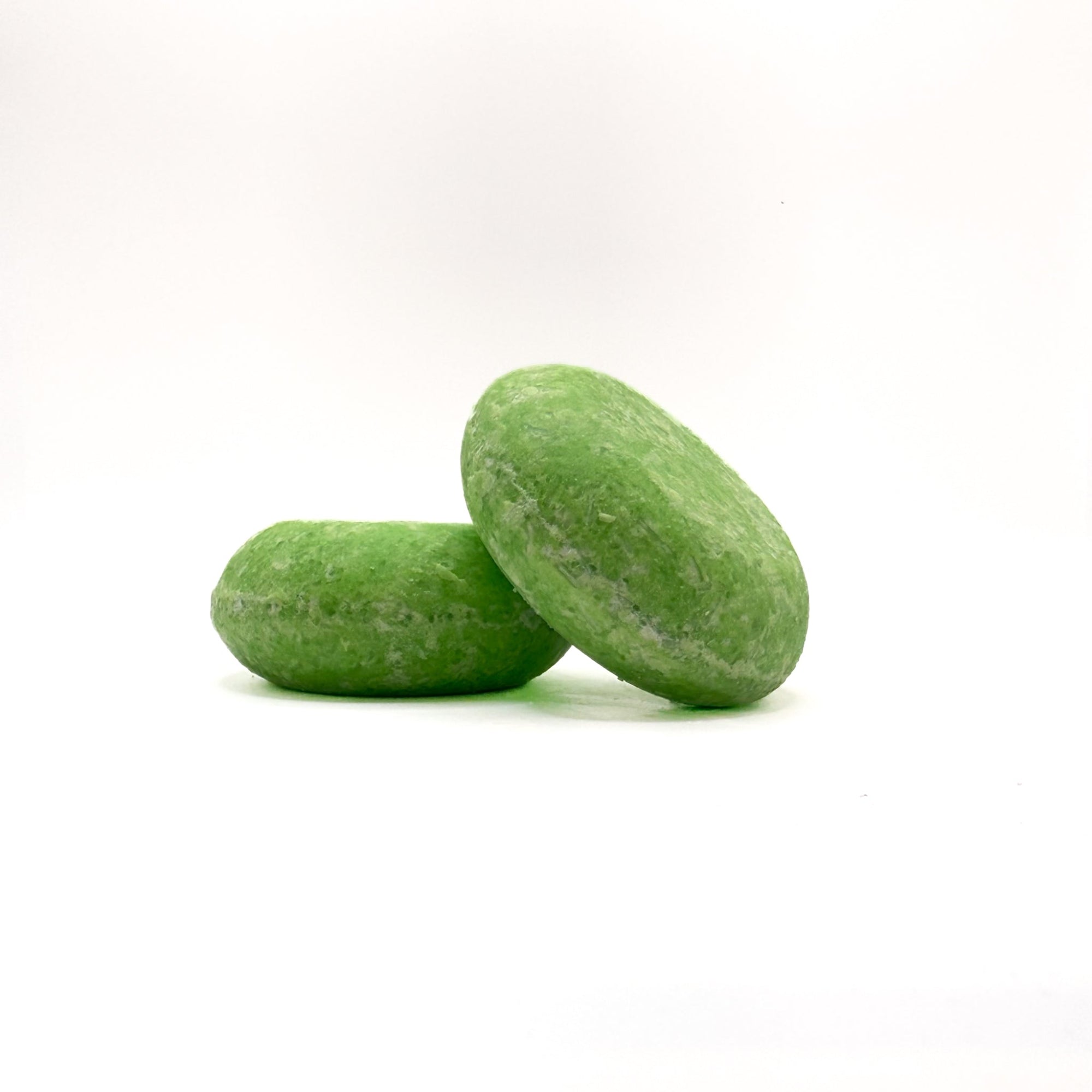 Two green round solid coconut lime cooler solid shampoo bars with white shavings from Rusted Acre Soap Company. One leaning on the other.