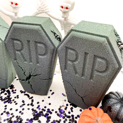 Two gray coffin shaped foam bath bombs with 'RIP' embossed on them, displayed on a white surface with Halloween props including small skeletons, purple glitter, and miniature pumpkins.
