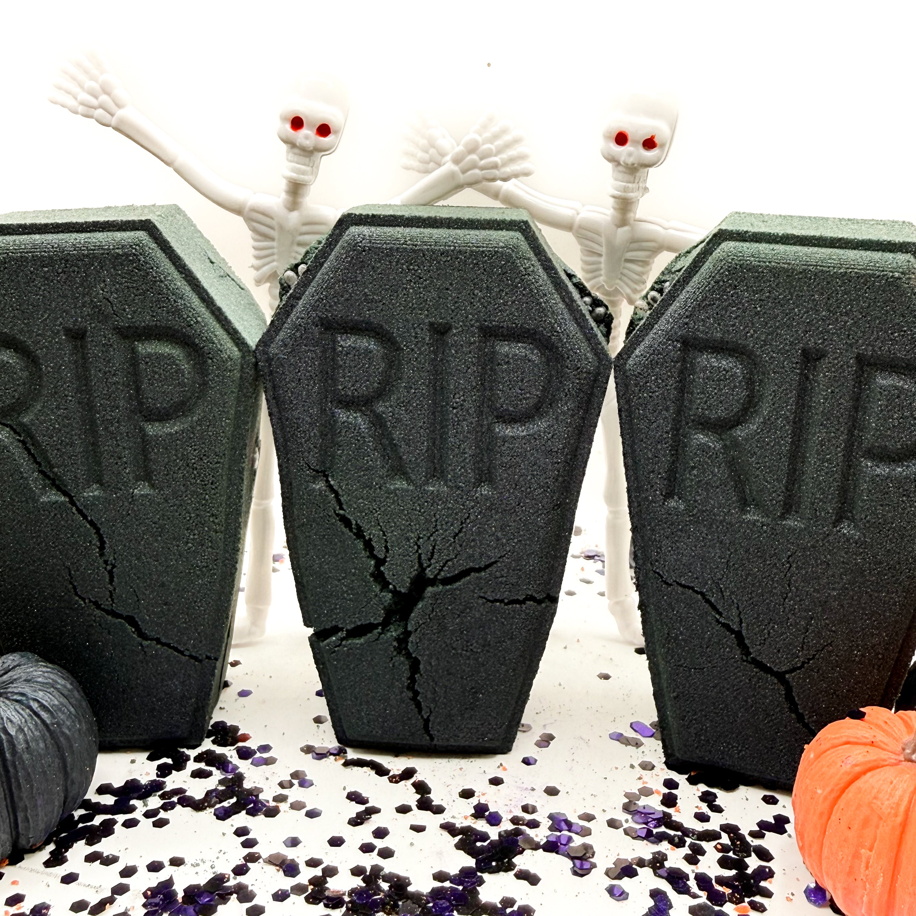 Three black coffin shaped bath bombs with 'RIP' embossed on the front, arranged on a white surface with scattered purple and black confetti. Two white skeleton figures stand behind them, and a small orange pumpkin is visible in the lower right corner.