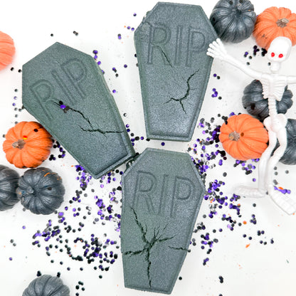 Three gray coffin shaped bath bombs with 'RIP' embossed on them, arranged on a white background with Halloween props including small pumpkins, black and purple glitter, and a white plastic skeleton.