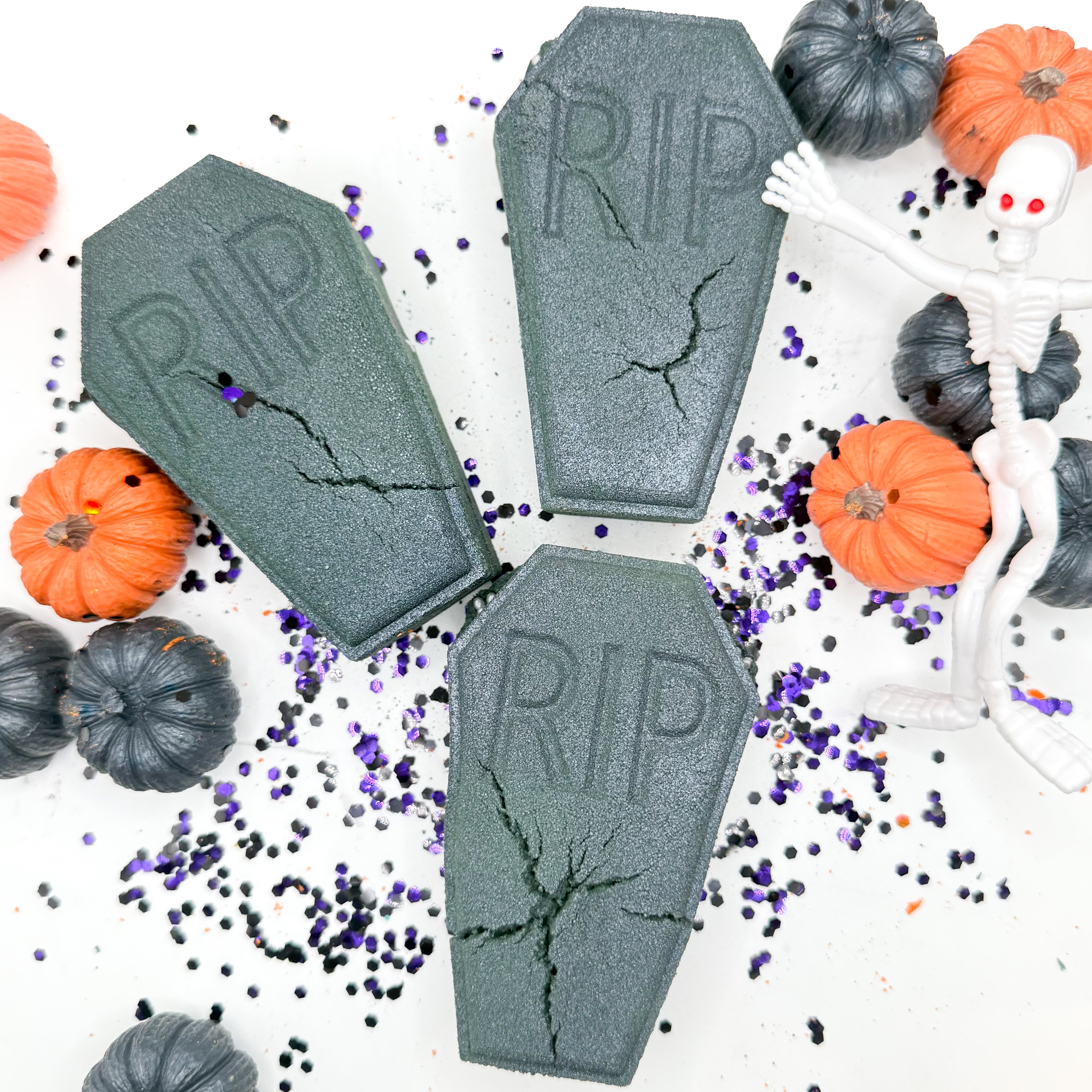 Three gray coffin shaped bath bombs with 'RIP' embossed on them, arranged on a white background with Halloween props including small pumpkins, black and purple glitter, and a white plastic skeleton.