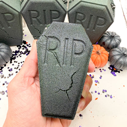 A hand holds a dark gray coffin shaped bath bomb with 'RIP' embossed on it, with more matching coffin-shaped items and small pumpkins in the background.