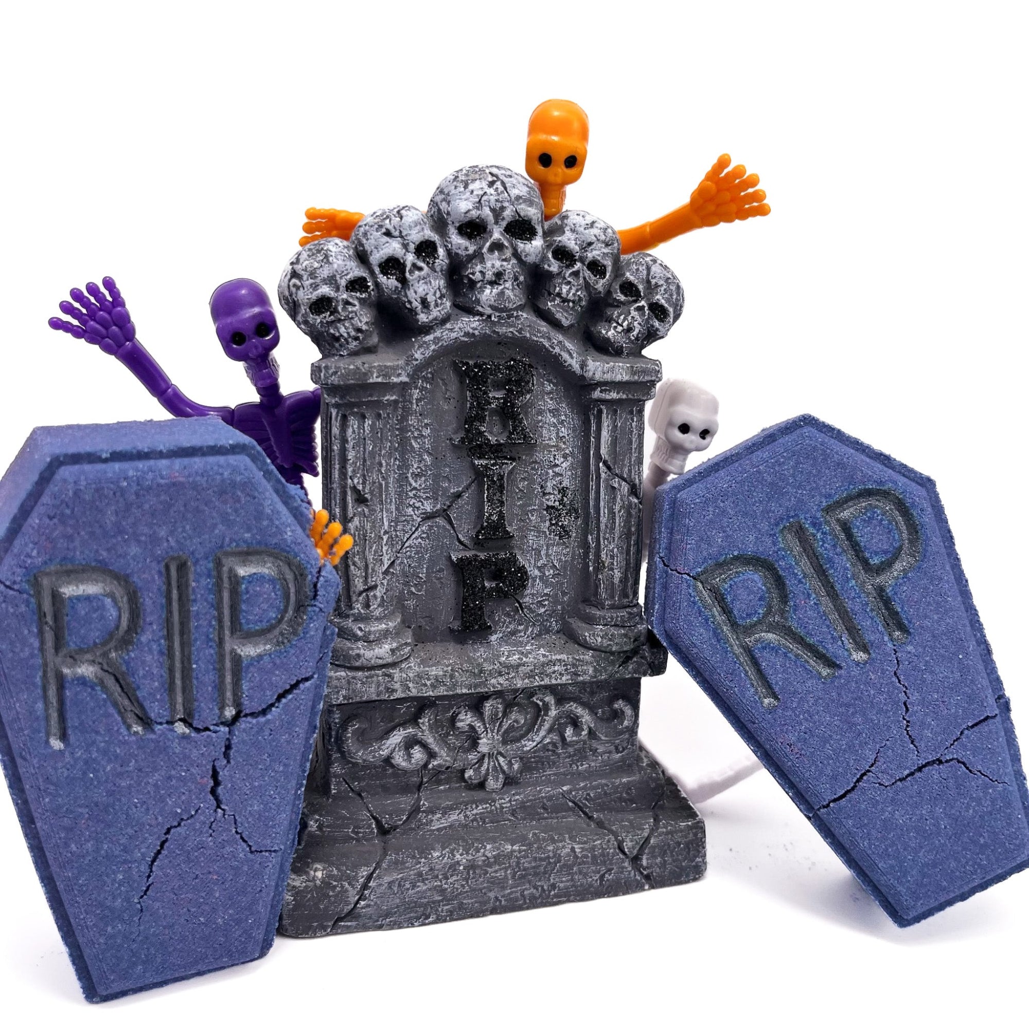 Two dark blue-purple coffin bath bombs from Rusted Acre Soap Co. Displayed with one purple and one orange rubber skeleton and an alter with skulls on top.