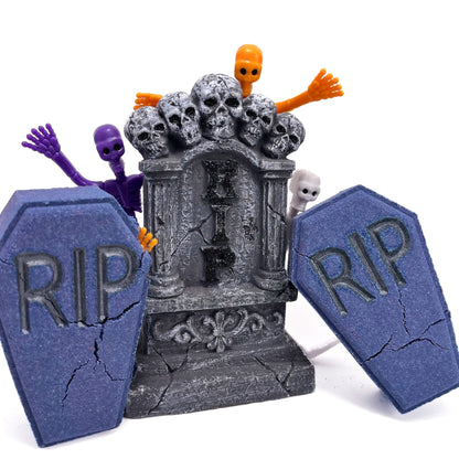 Two dark blue-purple coffin bath bombs from Rusted Acre Soap Co. Displayed with one purple and one orange rubber skeleton and an alter with skulls on top.