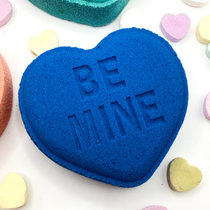 Blue heart shaped bath bomb with 'BE MINE' text on a white background with colorful heart-shaped candies.