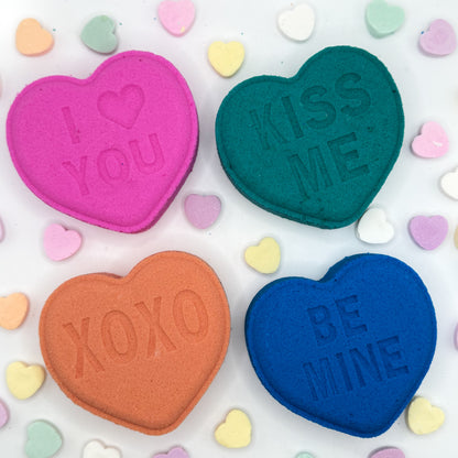 Four heart shaped bath bombs with messages 'I ❤️ YOU', 'KISS ME', 'XOXO', and 'BE MINE' on a colorful heart-patterned background.