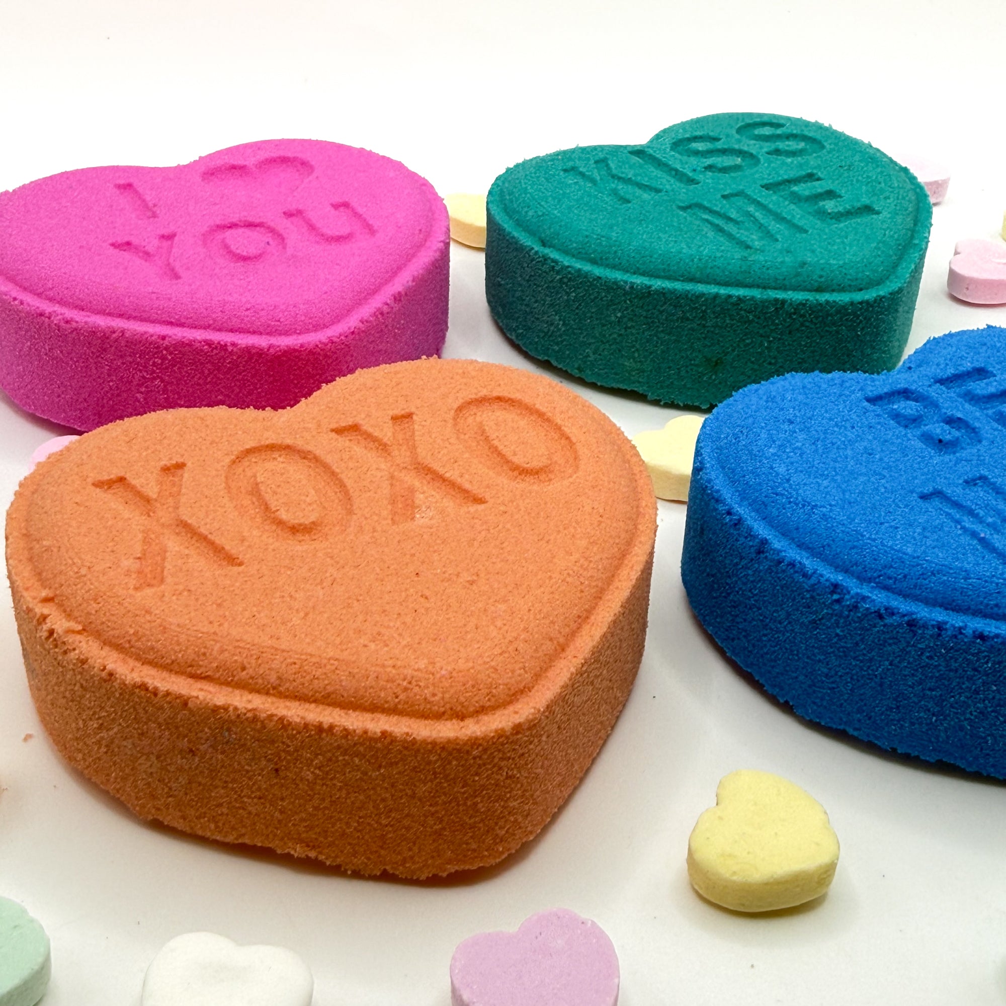 Four conversation heart inspired bath bombs laying flat in pink, peach, blue, teal on a white background.