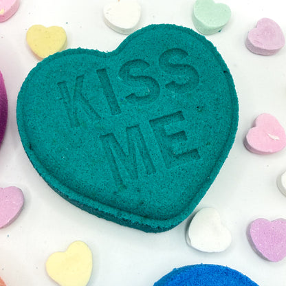 Teal heart shaped bath bomb with 'KISS ME' text on a white background with colorful heart-shaped candies.