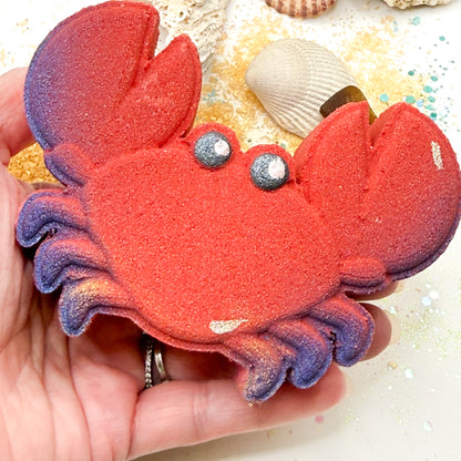 Red crab bath bomb with blue pinchers being held in a hand over a beach themed background. 